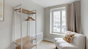 Una habitación moderna y minimalista con un sofá blanco, una estantería de madera y un amplio ventanal con cortinas.