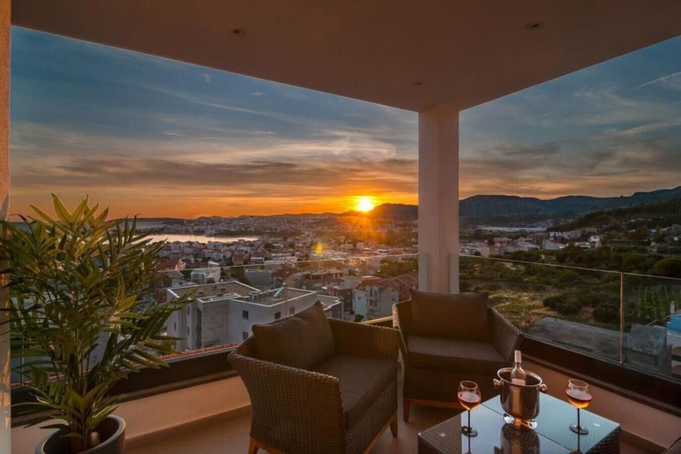 A balcony with a view of a sunset over a cityscape, furnished with a sofa and chairs, and a table with glasses and a bottle.