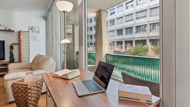 Ein Balkon mit Holztisch, Laptop und Büchern, mit Blick auf ein Gebäude und Grünflächen.