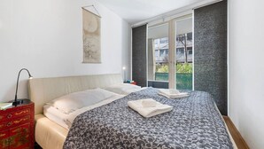 Ein Schlafzimmer mit Bett, Nachttisch, Lampe, einer roten Kommode, einem Wandbild und einem Fenster mit Blick auf Gebäude und Grünflächen.