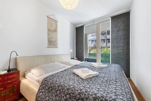 Ein Schlafzimmer mit Bett, Nachttisch, Lampe, einer roten Kommode, einem Wandbild und einem Fenster mit Blick auf Gebäude und Grünflächen.