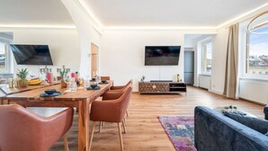 Ein modernes Wohnzimmer mit einem Holzesstisch, Ledersesseln, einem Flachbildfernseher und großen Fenstern.