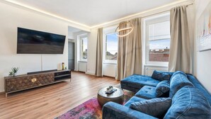 Ein modernes Wohnzimmer mit großem Fernseher, einem blauen Sofa, Holzboden und Blick auf die Stadt durch große Fenster.