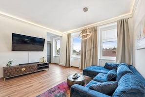 Ein modernes Wohnzimmer mit großem Fernseher, einem blauen Sofa, Holzboden und Blick auf die Stadt durch große Fenster.