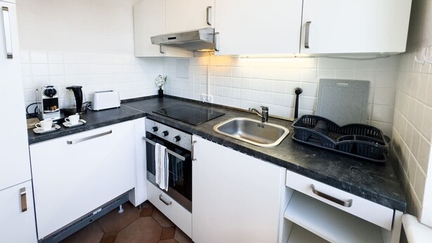 Eine kompakte Küche mit weißen Schränken, einer schwarzen Arbeitsplatte, einer Edelstahlspüle und einem eingebauten Backofen.