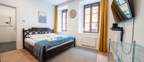 A bedroom with a bed, a clock, a window, a radiator, and a TV mounted on the wall.