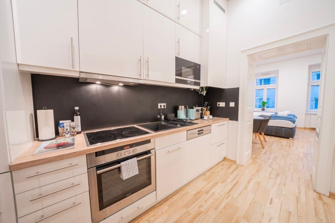 Een moderne keuken met een fornuis, oven en apparaten op het aanrecht. Door een open doorgang is een eethoek met een tafel en stoelen zichtbaar.