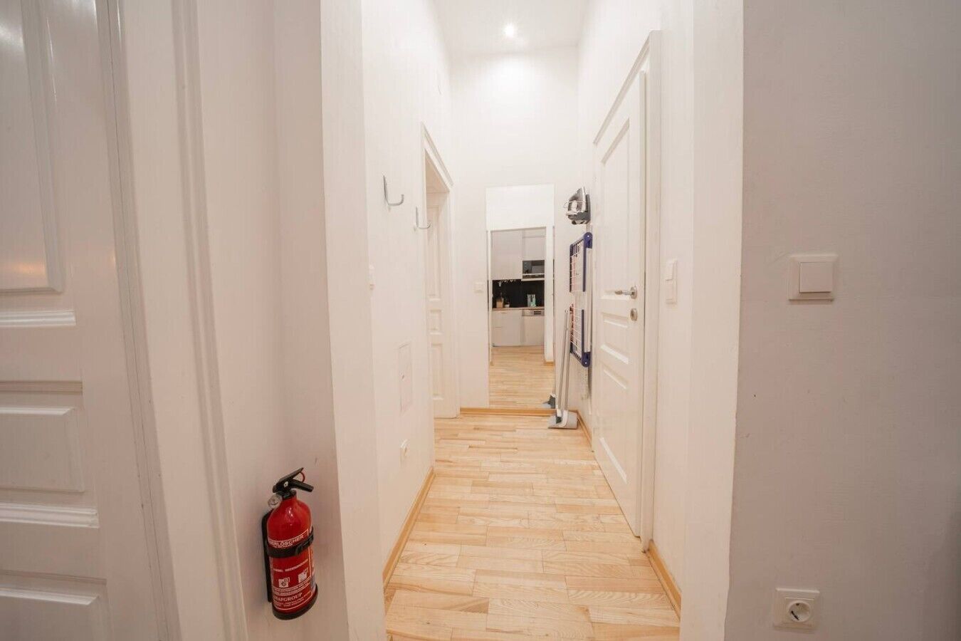 A hallway with white doors, a fire extinguisher, and a kitchen area in the distance.