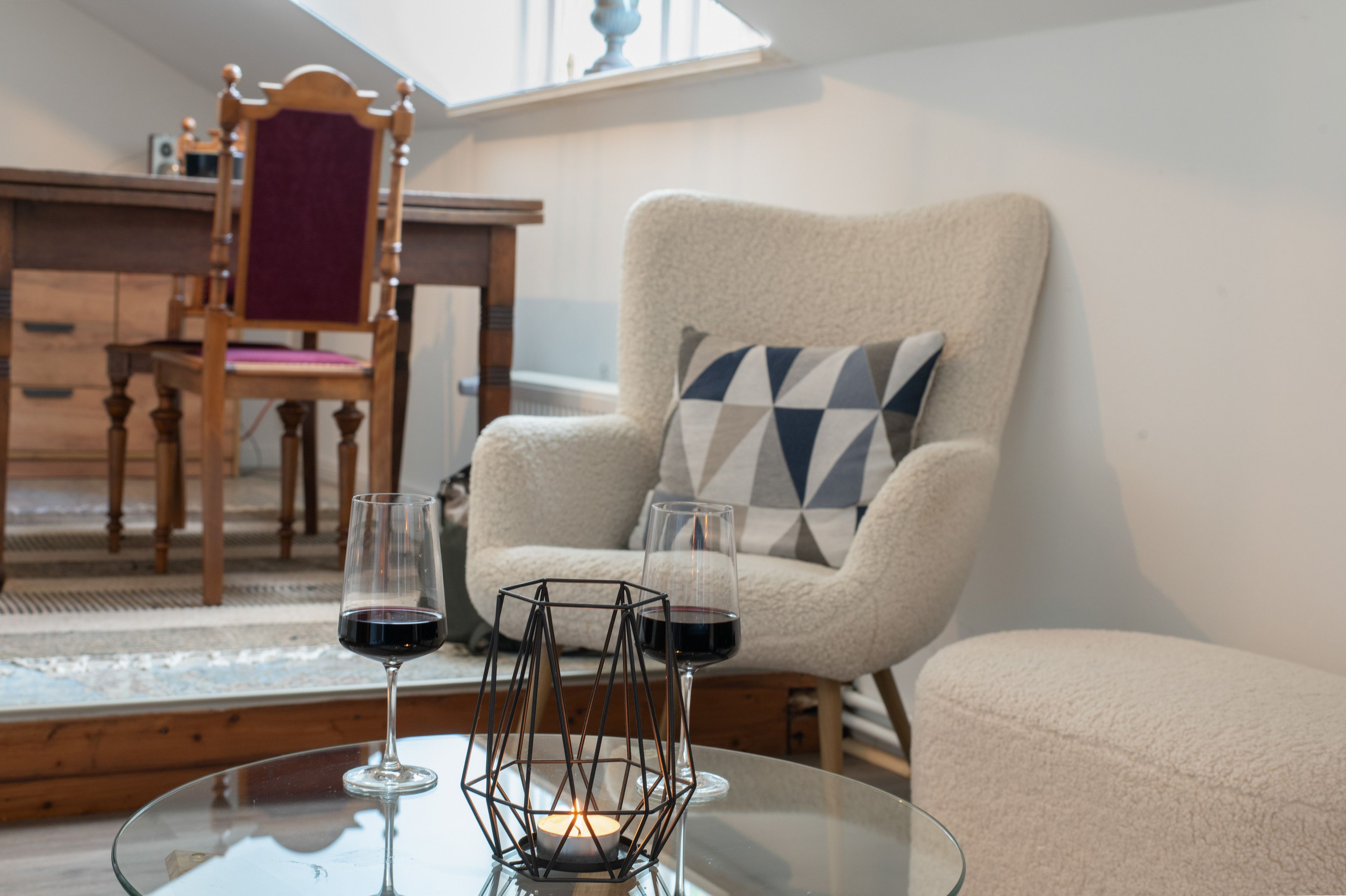 Een knus interieur met een comfortabele fauteuil, een glazen bijzettafeltje met twee wijnglazen en een kaars, en uitzicht op het omliggende landschap door een raam.