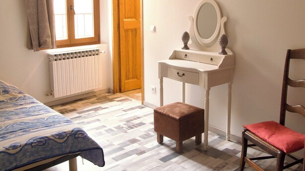 Een slaapkamer met een bed, een houten deur, een witte dressoir met spiegel, een rode stoel en een kamerkleed met dessin.