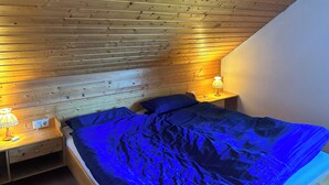 A bedroom with a sloped ceiling, wooden walls, a bed with blue bedding, two bedside tables with lamps, and a white wall on one side. - Große Ferienwohnung 1 Oberzarten im Gästehaus Lickert (Hinterzarten)