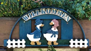 A wooden welcome sign with two duck figures, one in a blue outfit and the other in white, with a blue hat. - Große Ferienwohnung 1 Oberzarten im Gästehaus Lickert (Hinterzarten)
