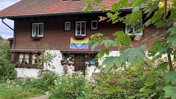 A two-story house with a red-tiled roof, a garden in front, and a rainbow flag hanging on the front door. - Große Ferienwohnung 1 Oberzarten im Gästehaus Lickert (Hinterzarten)