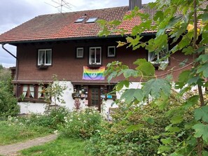 Ein zweistöckiges Haus mit rotem Ziegeldach, einem Vorgarten und einer Regenbogenflagge an der Haustür.