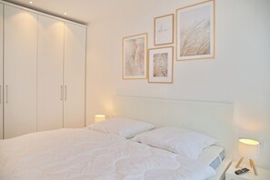 Une chambre à coucher avec un lit, du linge de lit blanc, une table de chevet avec une lampe et des tableaux encadrés au mur.