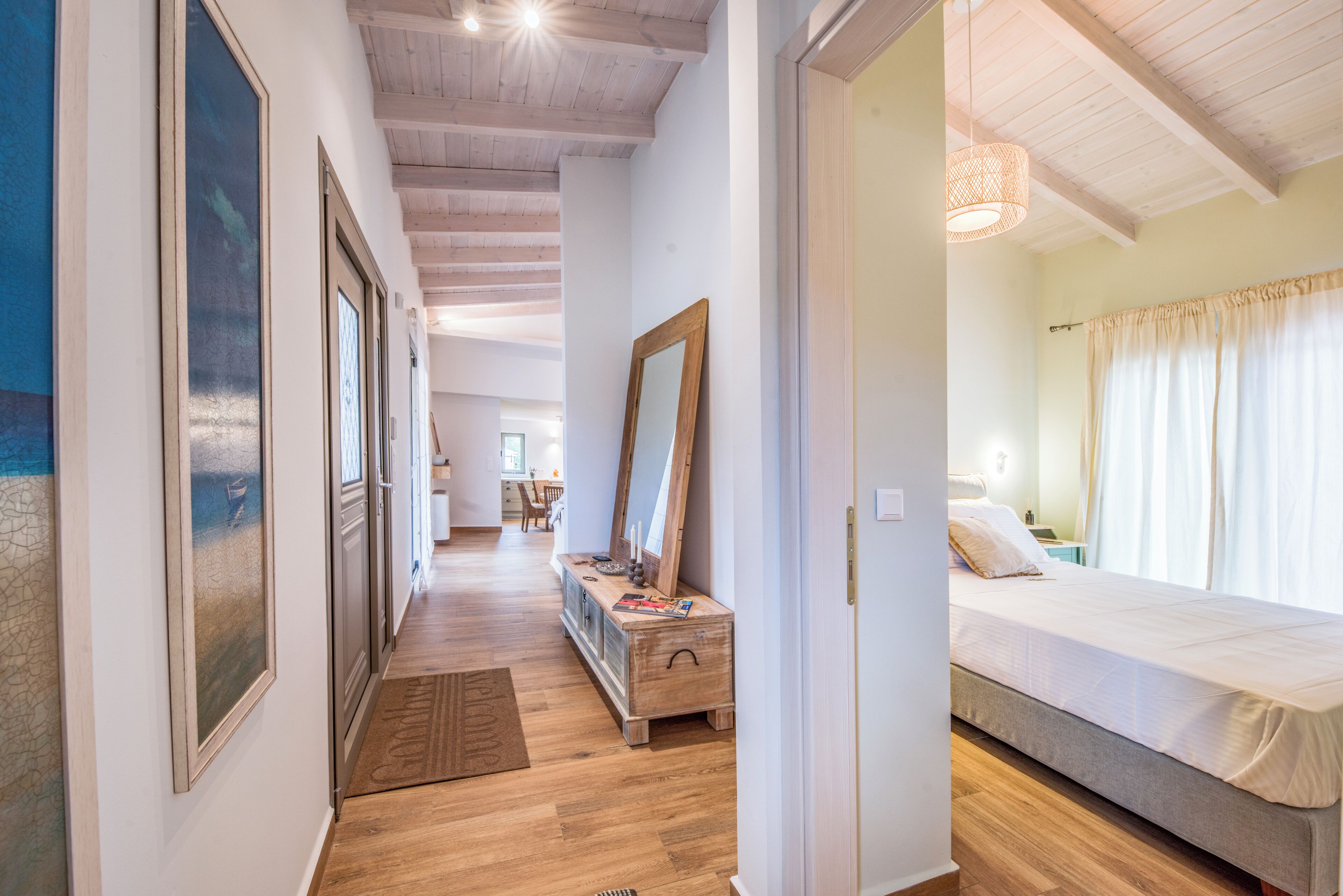 A hallway with wooden flooring, framed artwork on the walls, a wooden dresser, and a bed with a white bedspread in the adjacent room.