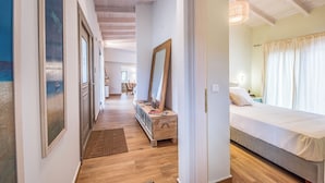 Ein Flur mit Holzboden, gerahmten Bildern an den Wänden, einer Holzkommode und einem Bett mit weißer Bettdecke im angrenzenden Zimmer.