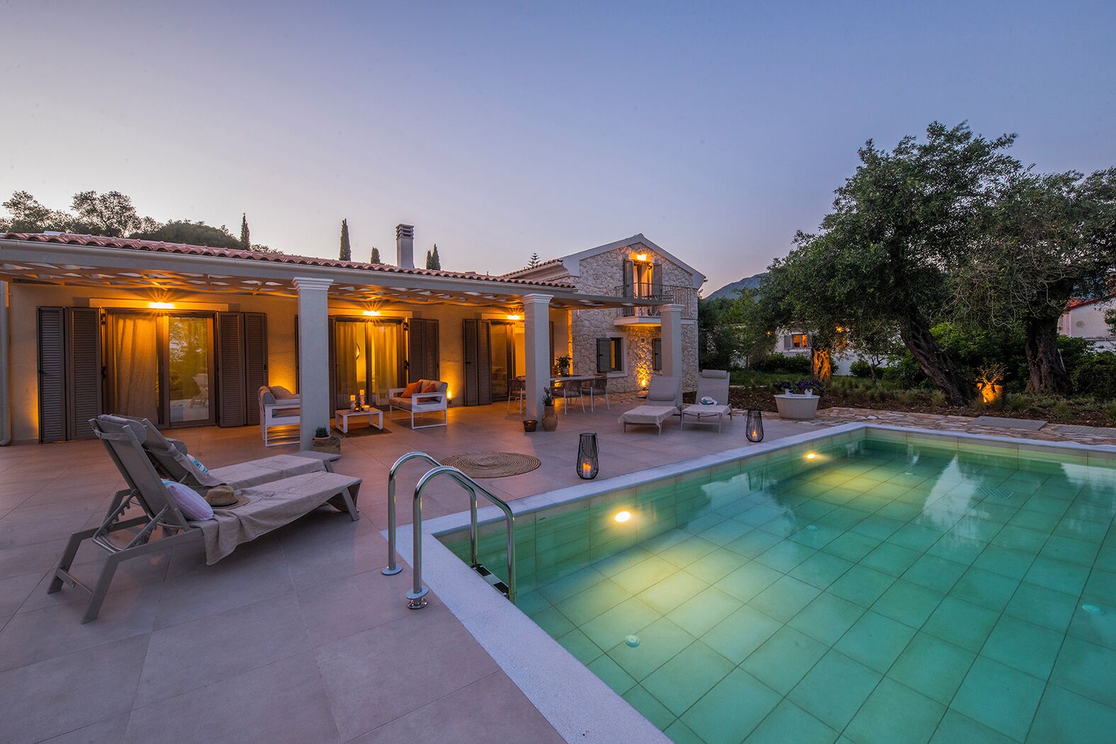 Ein Poolbereich mit Liegestühlen, ein Haus aus Stein mit einem Balkon und ein klares Schwimmbecken.