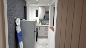 A narrow hallway leading to a kitchen with a refrigerator, a blender, and a window with blinds. - Agape Hubs (Barnstone)