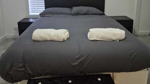 A modern bedroom with a large bed, two bedside tables, a window with blinds, and a black ottoman with decorative buttons. - Agape Hubs (Barnstone)