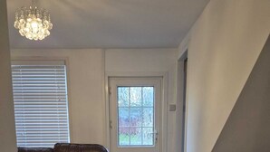 A living room with a brown leather sofa, a white door, a window with blinds, a chandelier, and a small heater. - Agape Hubs (Barnstone)