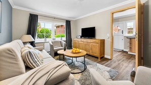 A modern living room with a sofa, a coffee table, a TV, and a kitchen area visible through an open door. - Tinkas Kate - Charming bungalow with garden in Westerland, Sylt (Sylt)