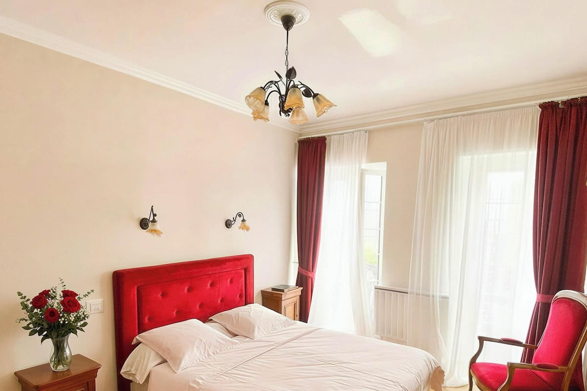 Une chambre avec un grand lit, une tête de lit rembourrée de couleur rouge, une table de chevet en bois ornée d’un vase de fleurs, un fauteuil rouge et un ventilateur de plafond.