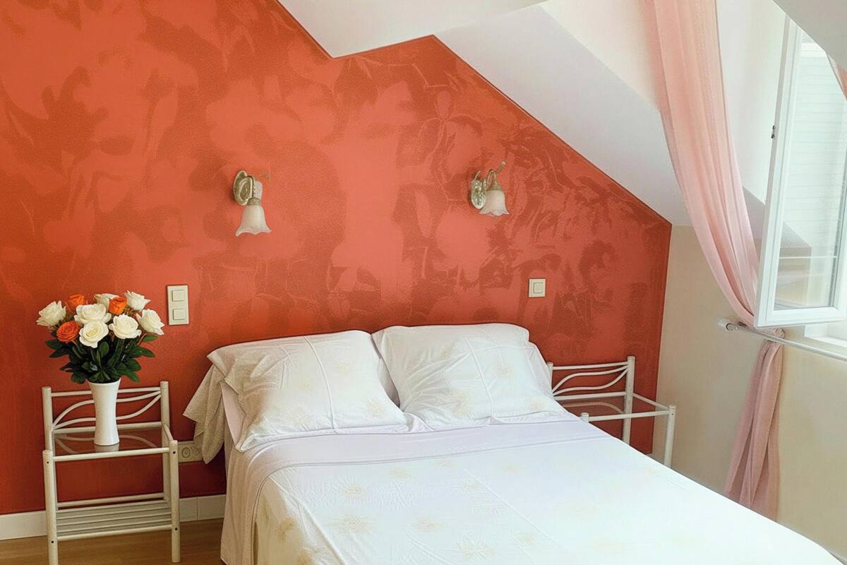 Une chambre avec un plafond en pente, un lit avec une literie blanche, une table de chevet ornée d’un vase de fleurs et une fenêtre avec des rideaux.