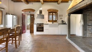 A kitchen with a central island, wooden chairs, a built-in oven, and a stone fireplace. - Il Focolare – Spacious Apartment in Farmhouse with Private Terrace and Pool (Pelago)