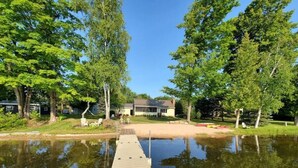 A wooden dock extending into a calm lake, surrounded by lush greenery and a house in the background. - Bass Bay Getaway | Lakefront Family Cottage (Elk Rapids)