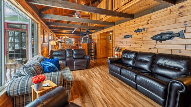 A cozy cabin interior with a wooden ceiling, a black leather sofa, a plaid sofa, and a dining area with a wooden table and chairs. - Bass Bay Getaway | Lakefront Family Cottage (Elk Rapids)