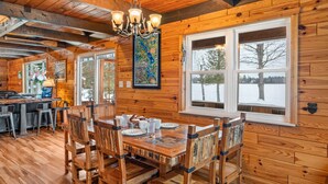 A wooden cabin interior with a dining area, large windows, and a chandelier. - Bass Bay Getaway | Lakefront Family Cottage (Elk Rapids)