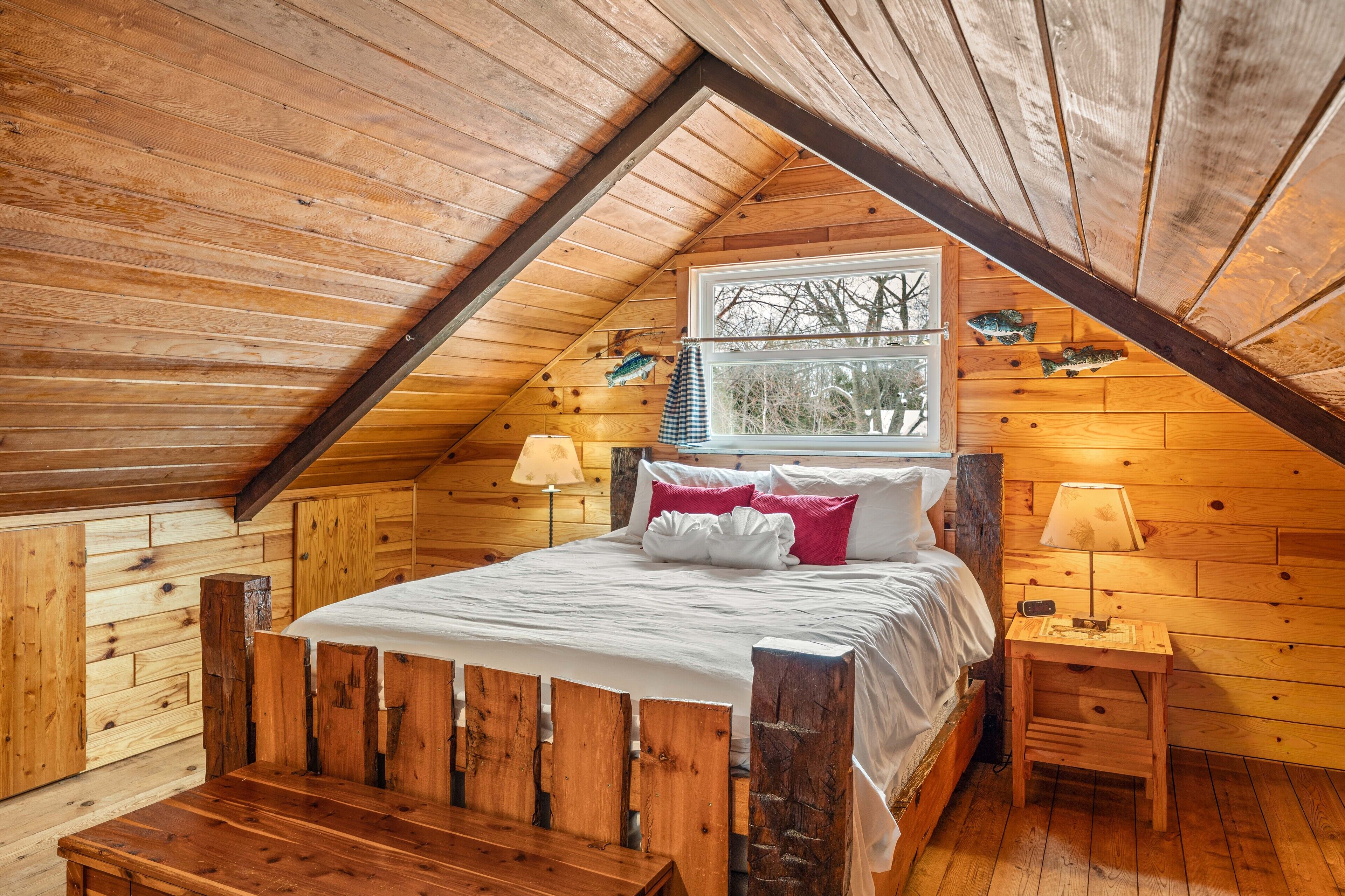 Een houten slaapkamer met schuine wanden, een bed met wit beddengoed, twee bedlampjes en een raam met uitzicht op bomen.