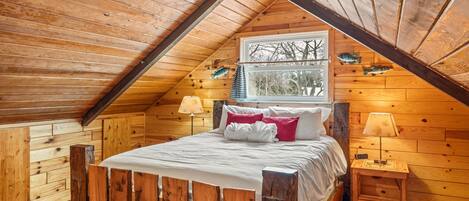 Een houten slaapkamer met schuine wanden, een bed met wit beddengoed, twee bedlampjes en een raam met uitzicht op bomen.