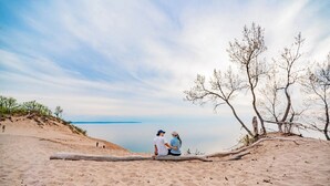 A sandy beach with a couple sitting on a log, overlooking a calm body of water and sparse trees. - Bass Bay Getaway | Lakefront Family Cottage (Elk Rapids)