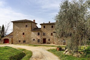 A stone building with multiple windows and a red door, surrounded by greenery and a gravel path. - La Torre – Panoramic Apartment in Farmhouse with Pool (Pelago)