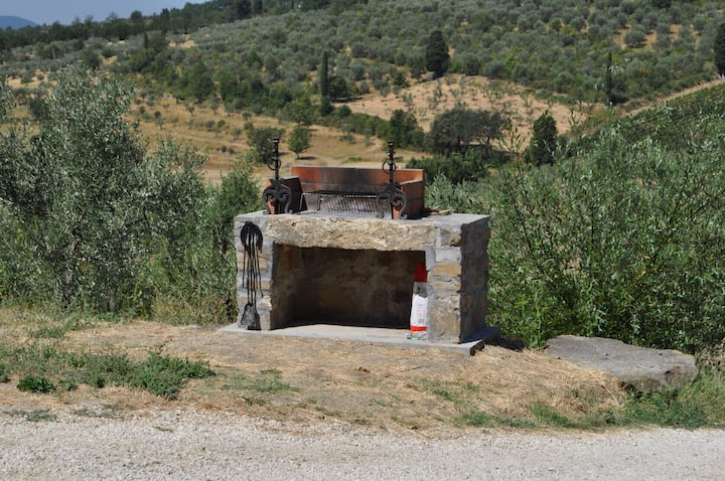 Une structure en pierre avec un bassin en métal et un banc en pierre, entourée d’oliviers et d’herbes sèches.