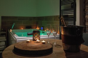 Une table en bois, ornée de bougies, de verres et d’une bouteille de champagne, se trouve dans une pièce où un jacuzzi est illuminé de lumière verte et où l’un des murs est en briques.