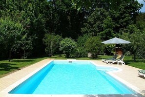 A rectangular swimming pool with a tiled edge, surrounded by a wooden deck. There are two lounge chairs and a single umbrella.