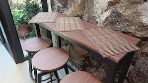 A bar area with a wooden table and stools, a stone wall, and a view of greenery outside.