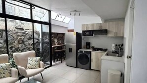 A modern kitchen with a dishwasher, washing machine, and a small sink. There is a glass table with chairs and a view of a stone wall.