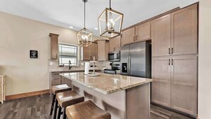 A modern kitchen with wooden cabinets, a stainless steel refrigerator, a microwave, and a granite countertop. There are three wooden bar stools and pendant lights above the island. - Hooked on Branson Cove - Pool Hot Tub Fireplace (Hollister)