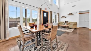 Uma sala de estar espaçosa com teto alto, grandes janelas, área de jantar com mesa de madeira e cadeiras de fibra, um sofá e uma pequena mesa de centro.