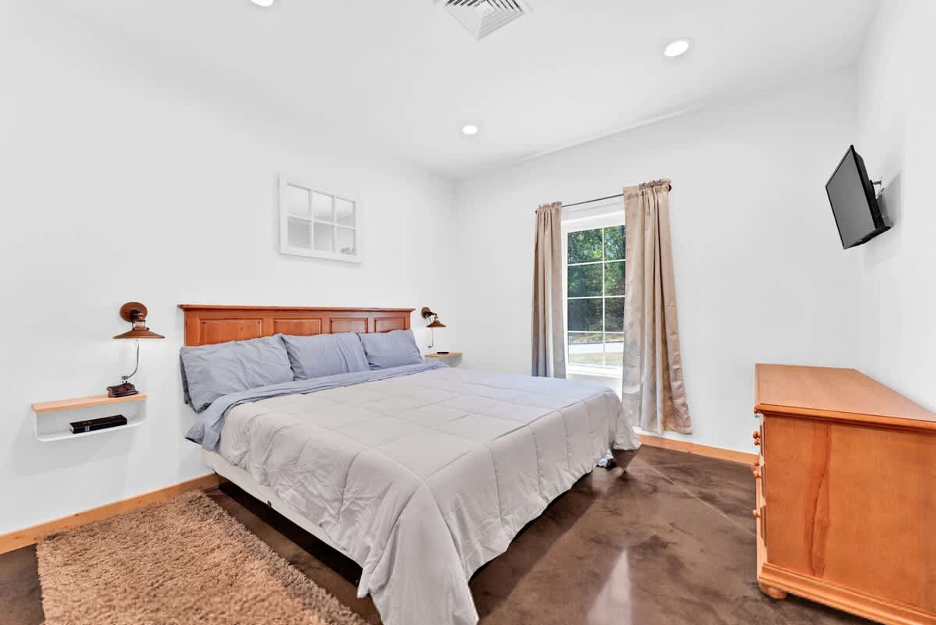 A bedroom with a bed, a TV mounted on the wall, a window with curtains, and a wooden nightstand with a lamp.