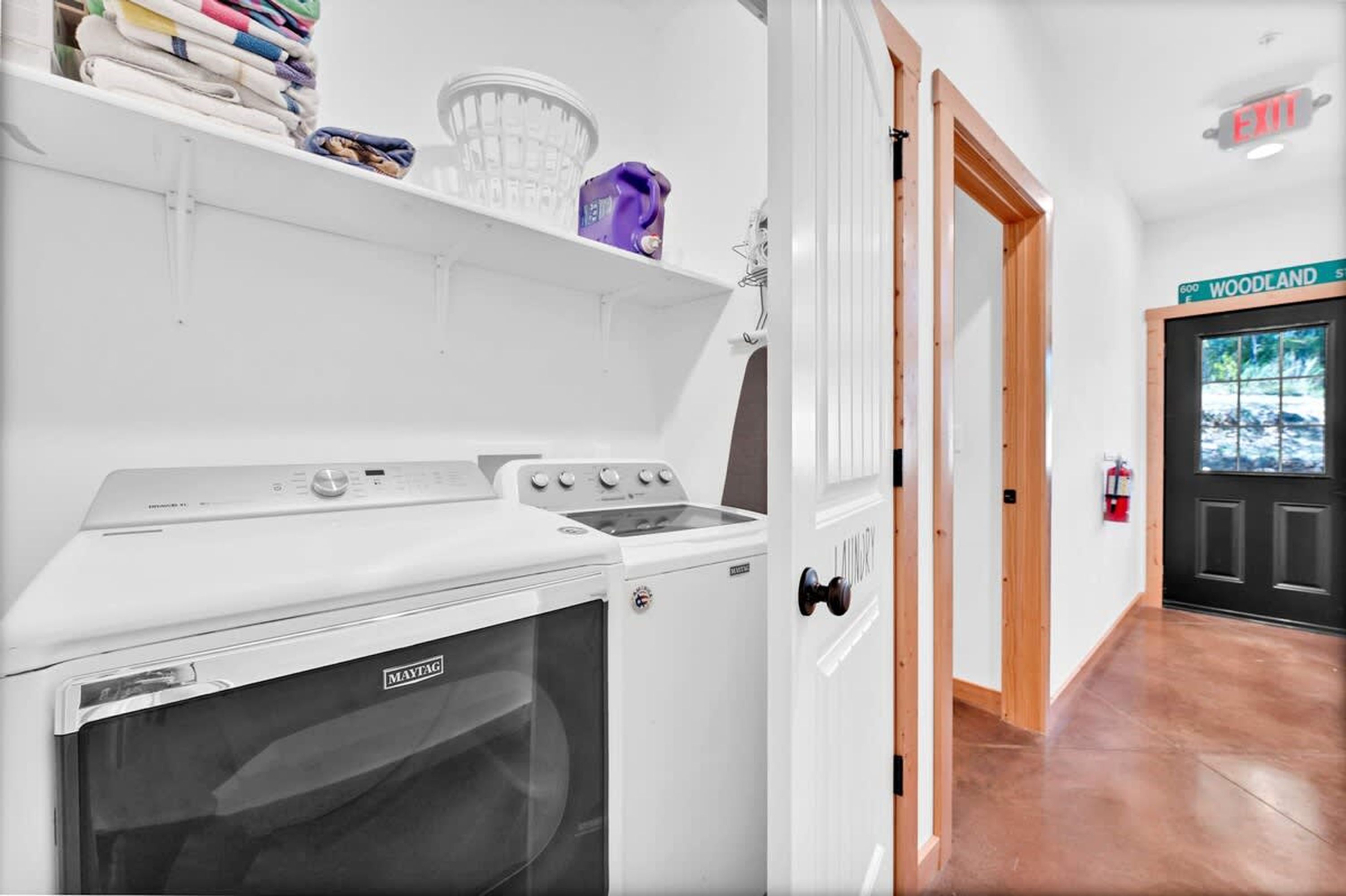 Une buanderie équipée d’un lave-linge et d’un sèche-linge Maytag, d’une étagère avec des vêtements pliés, et d’une porte donnant sur un couloir où est affiché un panneau indiquant « WOODLAND ».