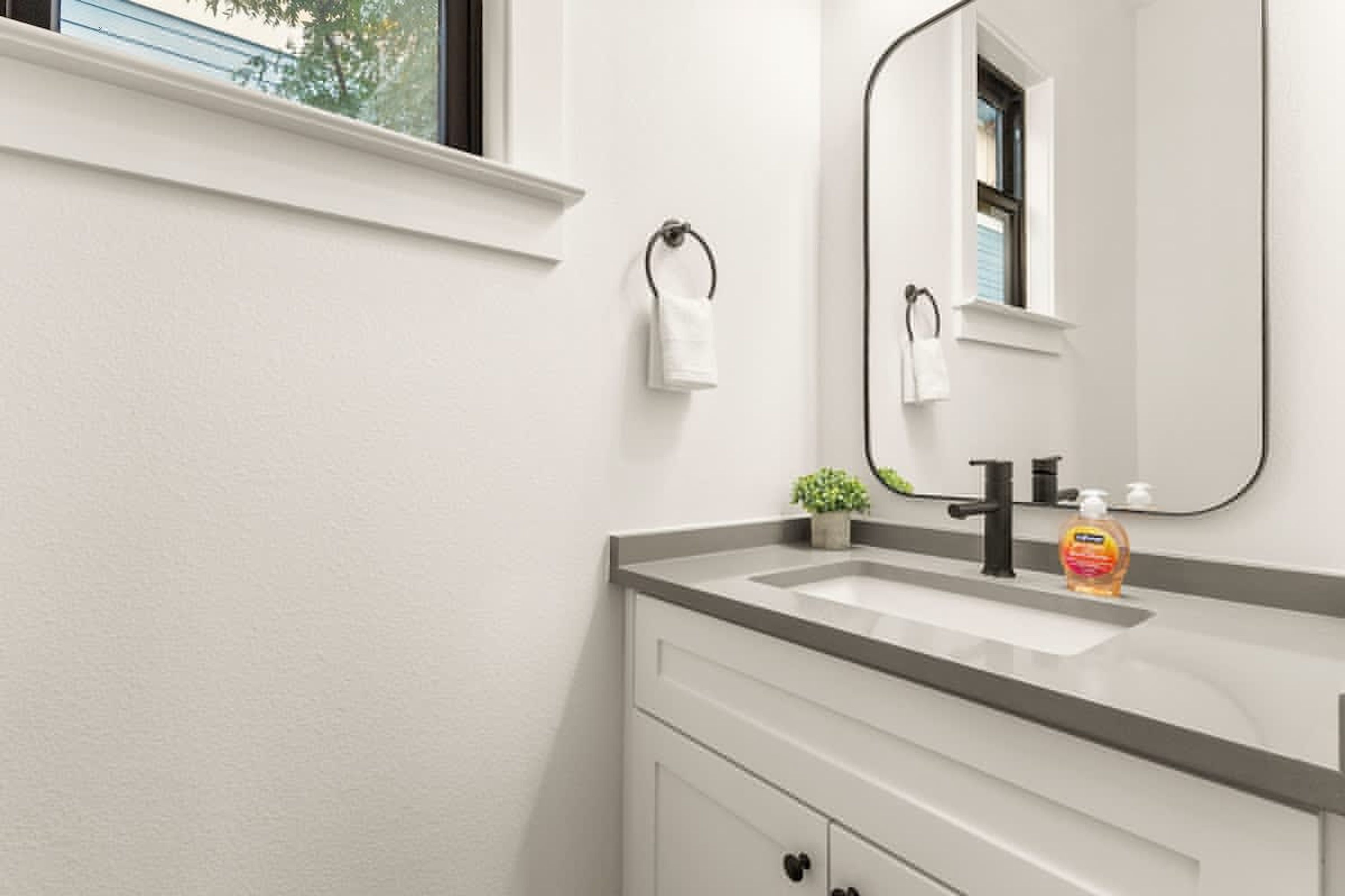 A bathroom with a white vanity, a black faucet, a mirror, and a window with a towel hanging on the wall.