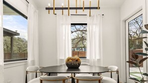 A dining area with a round table, six chairs, a modern chandelier, and large windows offering a view of the outdoors. - Austin Family Retreat at Ember House (Austin)