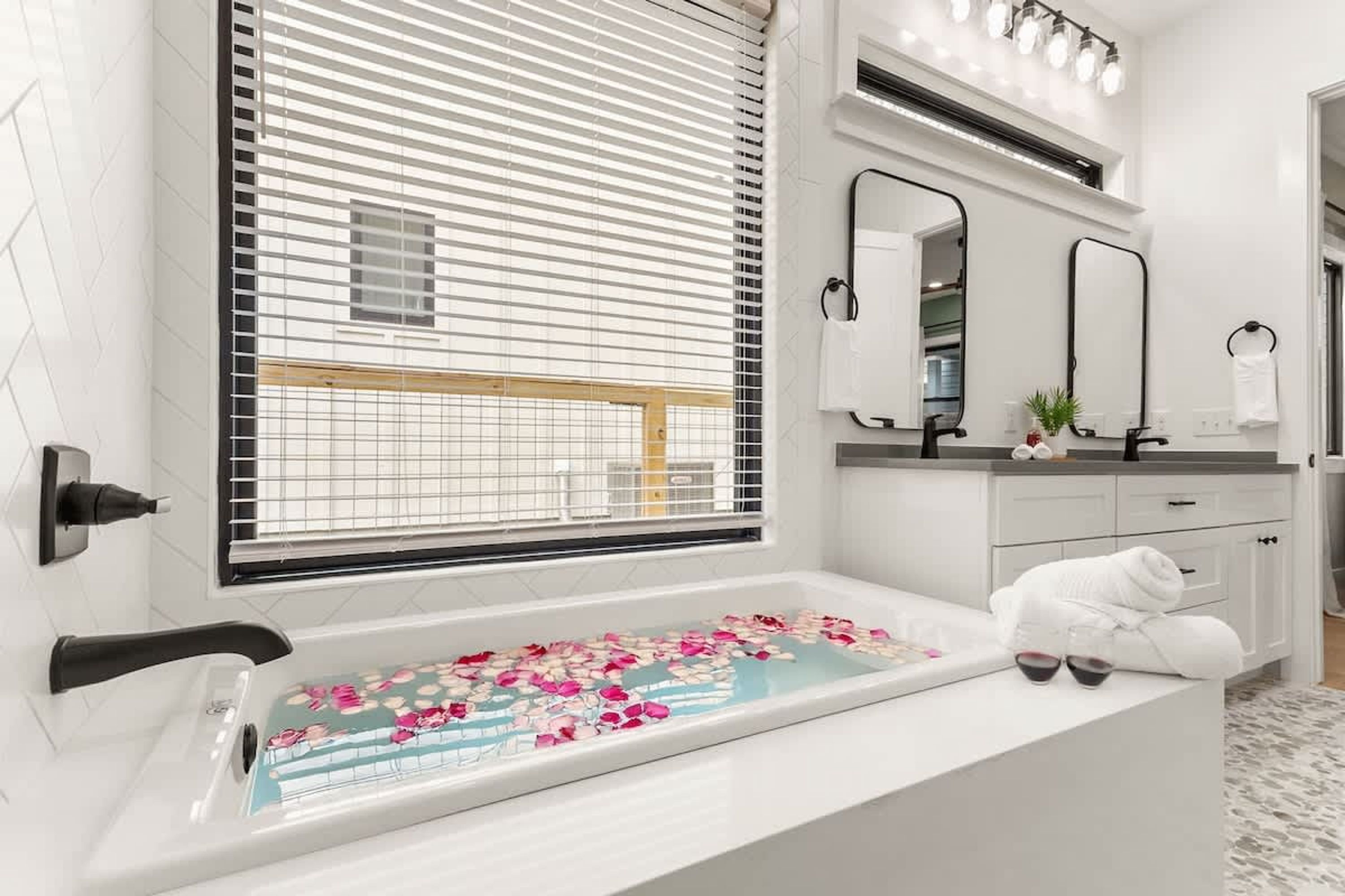 Een moderne badkamer met een badkuip, witte tegels, een raam met zonwering en een spiegel boven de wastafel.