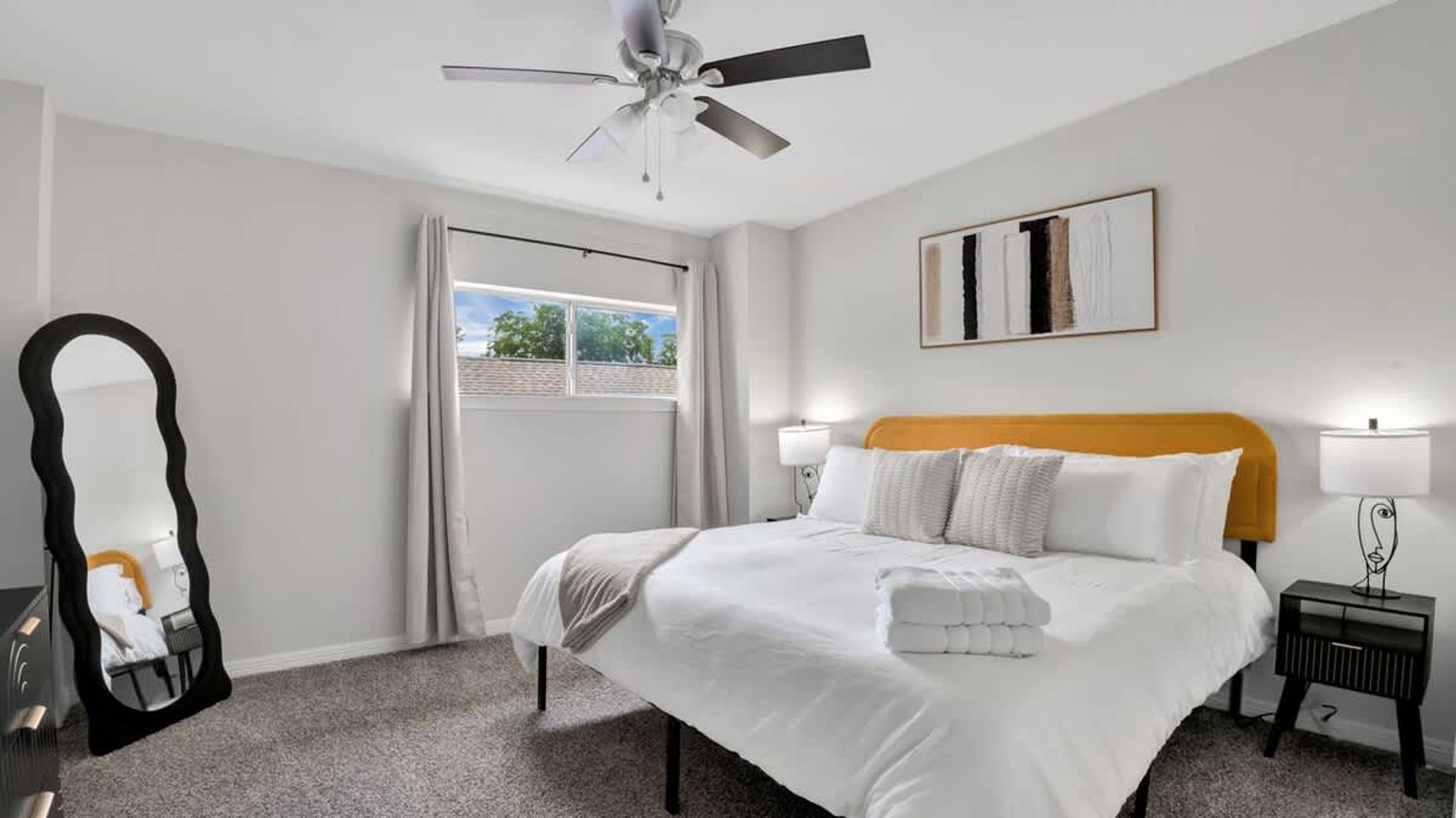 Une chambre d’hôtel avec un lit, un ventilateur de plafond, une fenêtre avec des rideaux, une œuvre d’art encadrée au mur et une table de chevet avec une lampe.