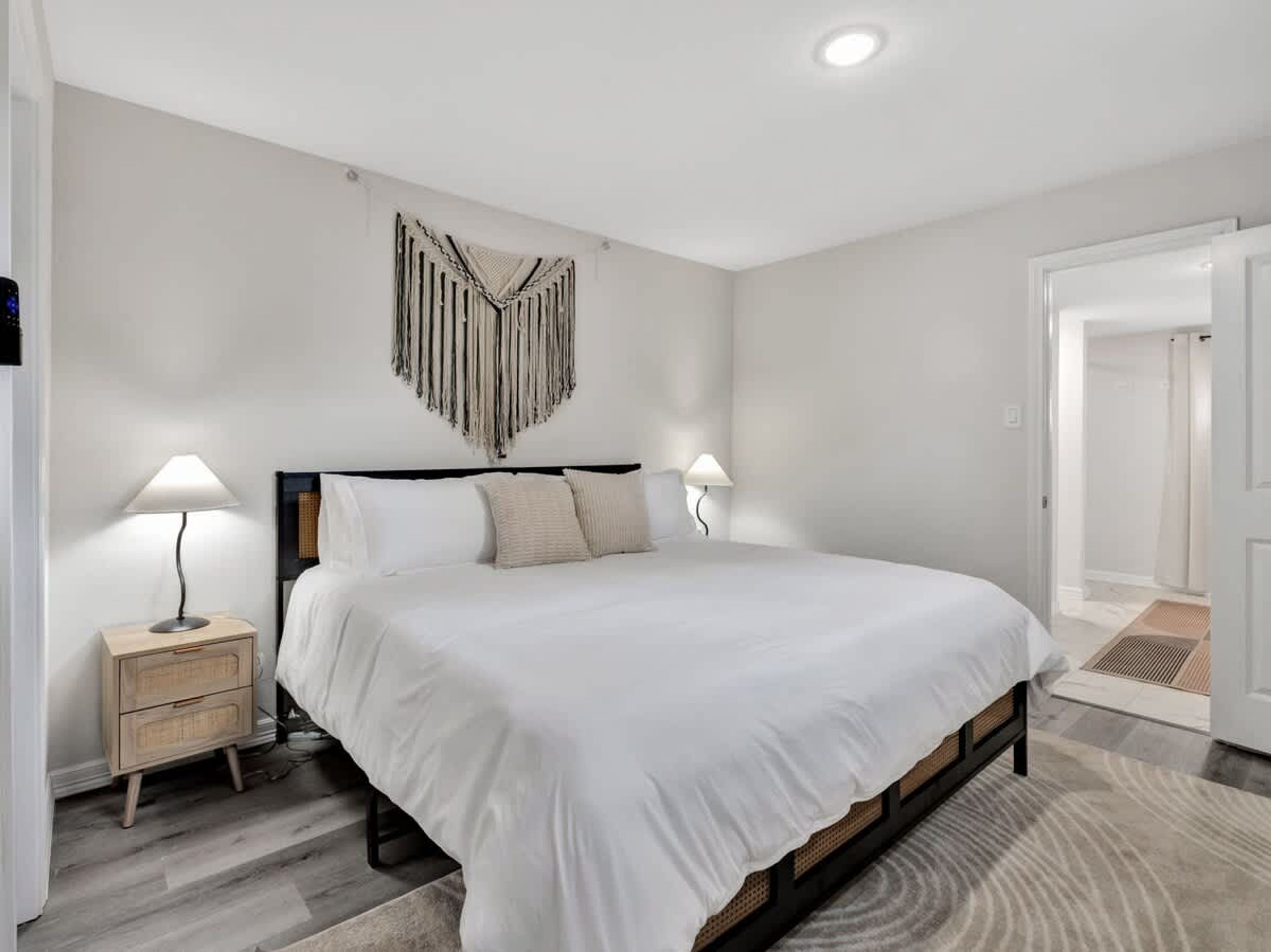 A bedroom with a large bed, two bedside tables with lamps, a wall-mounted macrame piece, and a door leading to another room.